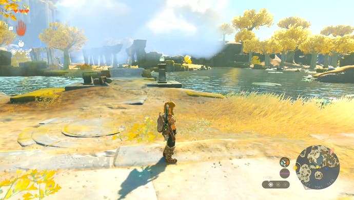 Link standing in an area with golden grass, trees with golden leaves as well as stone structures and a nearby lake in The Legend of Zelda: Tears of the Kingdom.